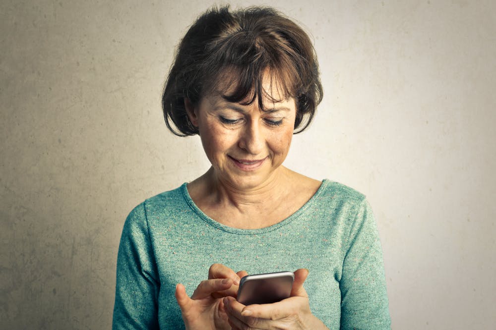 Mulher idosa sorridente, segurando celular cinza. Ilustração do texto sobre fisioterapia.