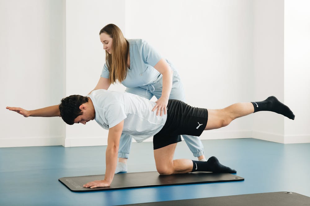Fisioterapeuta atenta, usando uniforme azul, acompanhando aluno em alongamento. Ilustração do texto sobre pilates para atletas.