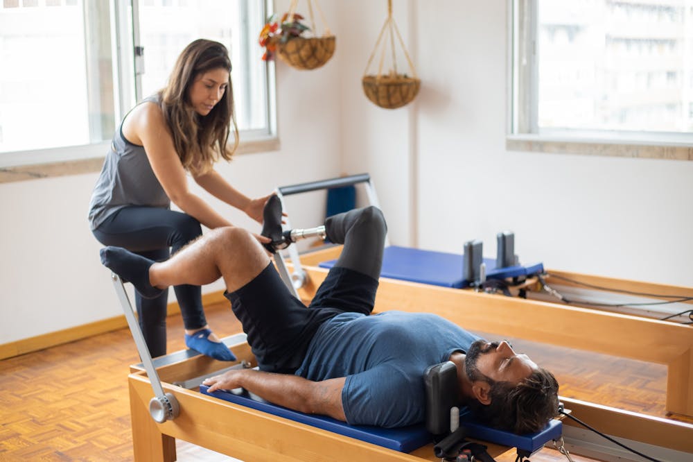 Homem usando blusa azul fazendo pilates com apoio de um aparelho. Ele tem uma perna protética e está sendo acompanhando por uma profissional. Ilustração do texto sobre pilates para atletas.
