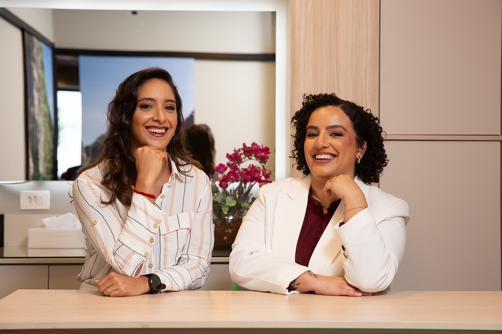 Dra. Carol e Dra. Stephany sorridentes sentadas próximas a uma bancada bege. Ilustração do texto sobre fisioterapia e menopausa.